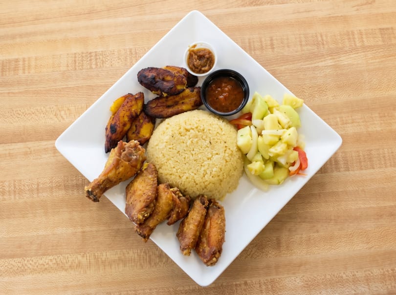 A white rectangular plate holds yellow rice, fried plantains, chicken wings, and a side salad.