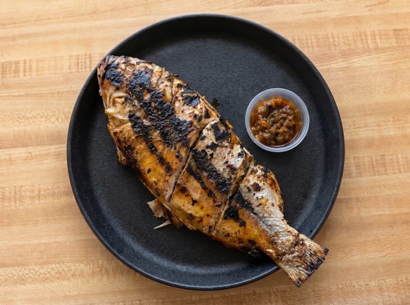 Grilled whole fish with dark char marks, served on a black plate with a side of sauce.