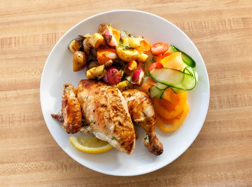Roasted chicken with potatoes, radishes, and a side salad of tomatoes, cucumbers, and yellow bell peppers.