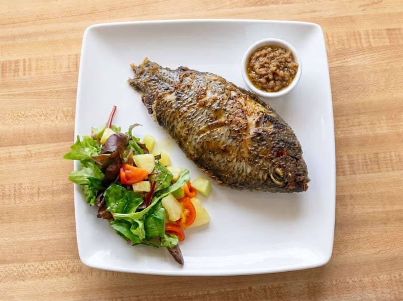 Whole grilled fish with a side salad of greens, tomatoes, and potatoes, and a small bowl of sauce.