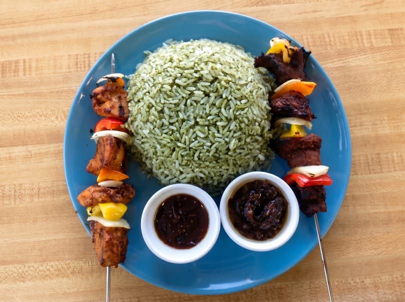 Two skewers of grilled meat and vegetables, green rice, and two dipping sauces on a blue plate.