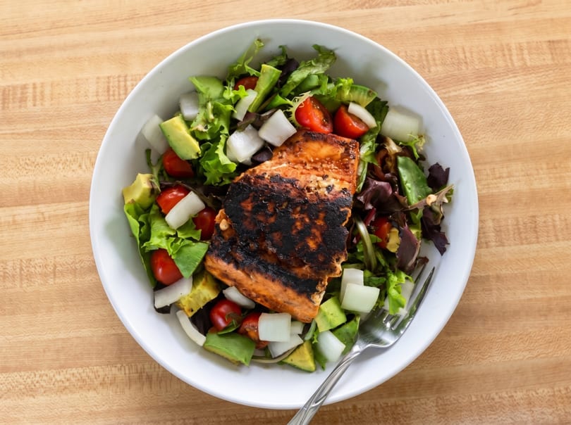 Grilled salmon salad with mixed greens, avocado, cherry tomatoes, and onions in a white bowl.