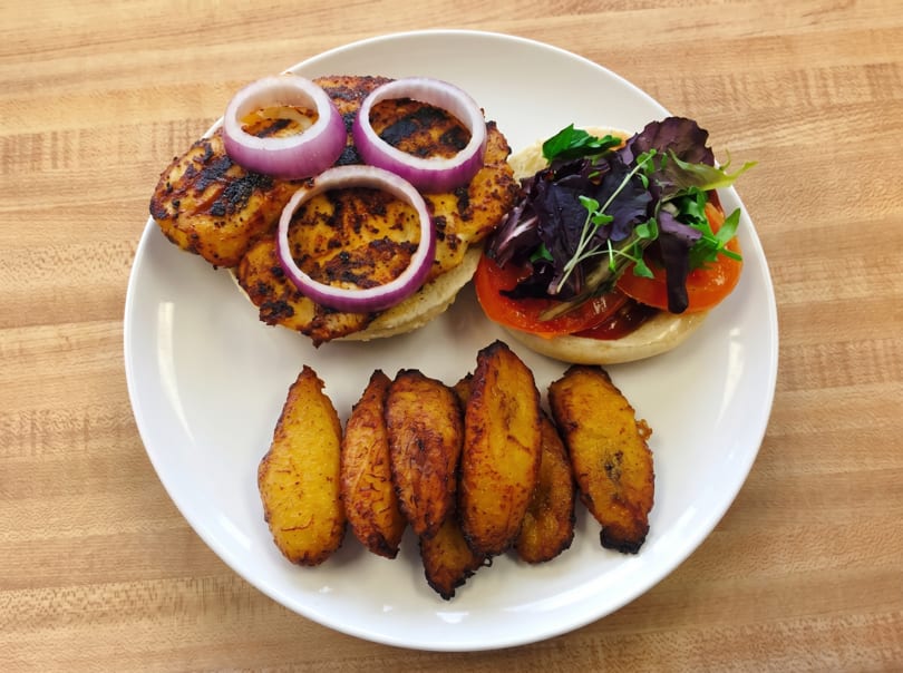 Grilled chicken sandwich with red onion, tomato, mixed greens, and a side of fried plantains.