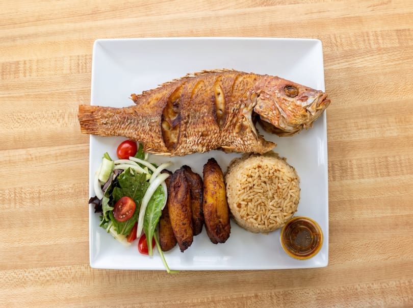 A whole fried fish with rice, plantains, and a side salad on a white rectangular plate.
