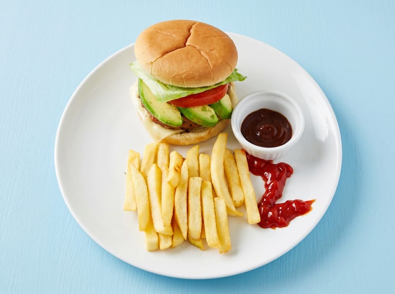 A burger with avocado, lettuce, and tomato, served with fries and ketchup on a white plate.