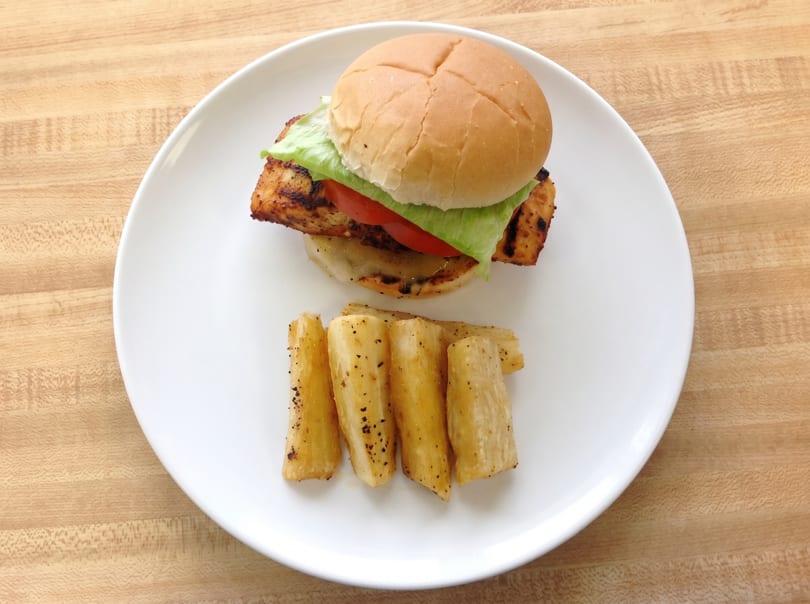 Grilled chicken sandwich with lettuce and tomato, served with seasoned yucca fries on a white plate.