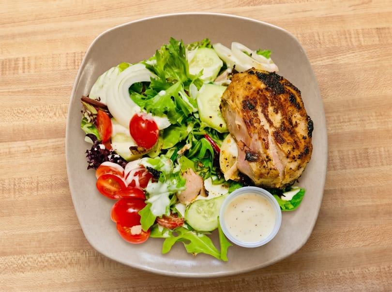Grilled chicken breast with a fresh garden salad, cherry tomatoes, cucumbers, and dressing on a plate.